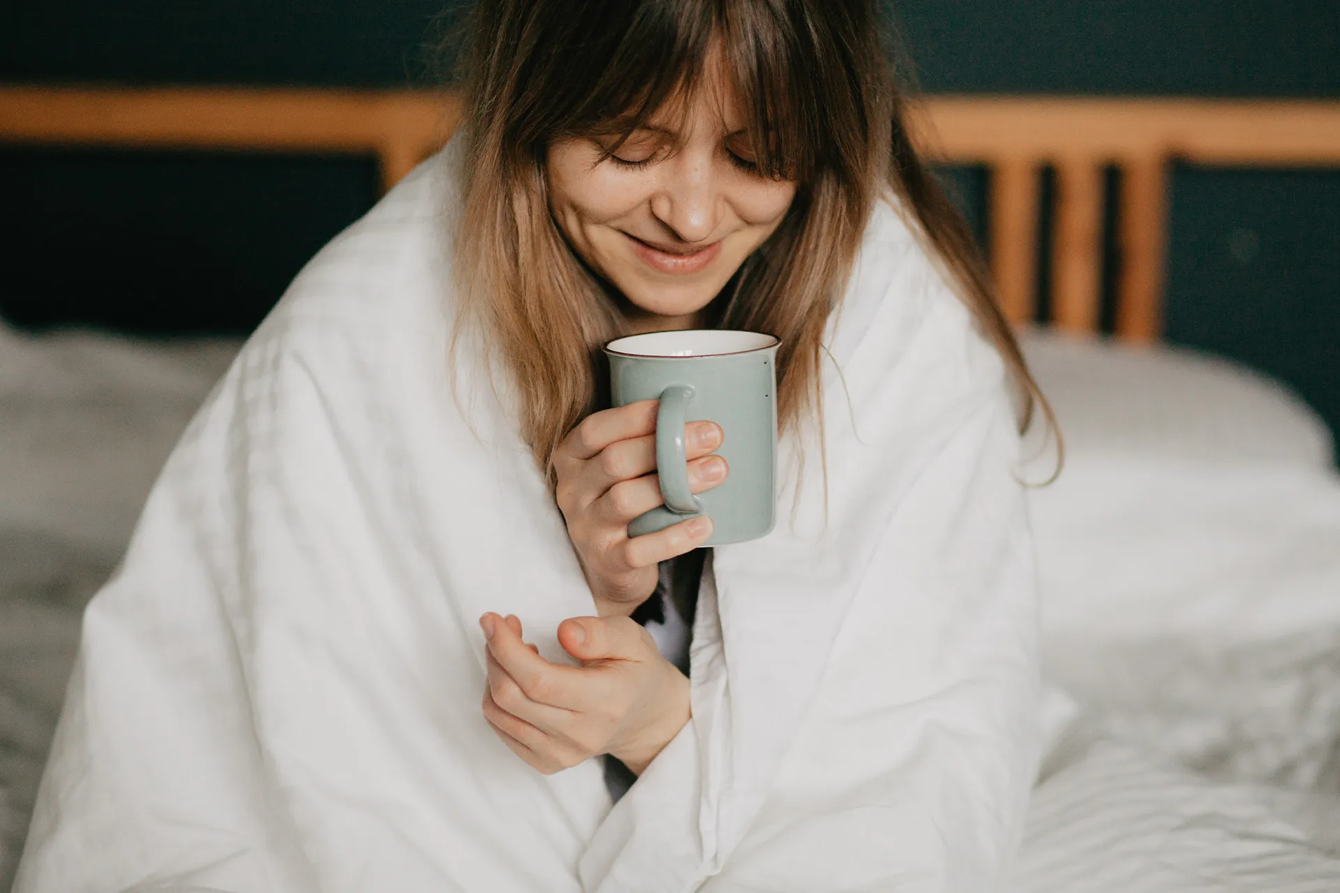 Frau sitzt in eine weiße Decke gehüllt im Bett und hält eine Tasse Kräutertee – Wärme und Ruhe zum Regelschmerzen lindern