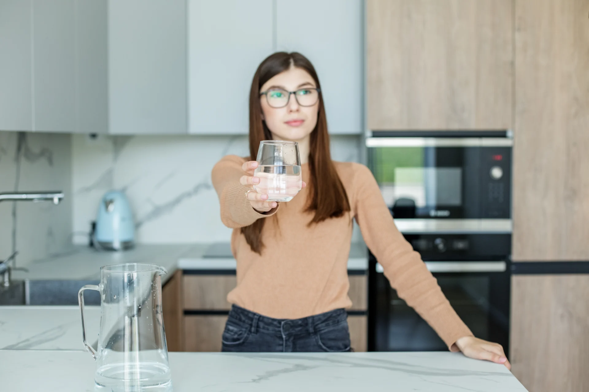 Junge Frau steht in der Küche und hält ein Glas Wasser in die Kamera – ausreichend trinken, um einer Blasenentzündung vorzubeugen