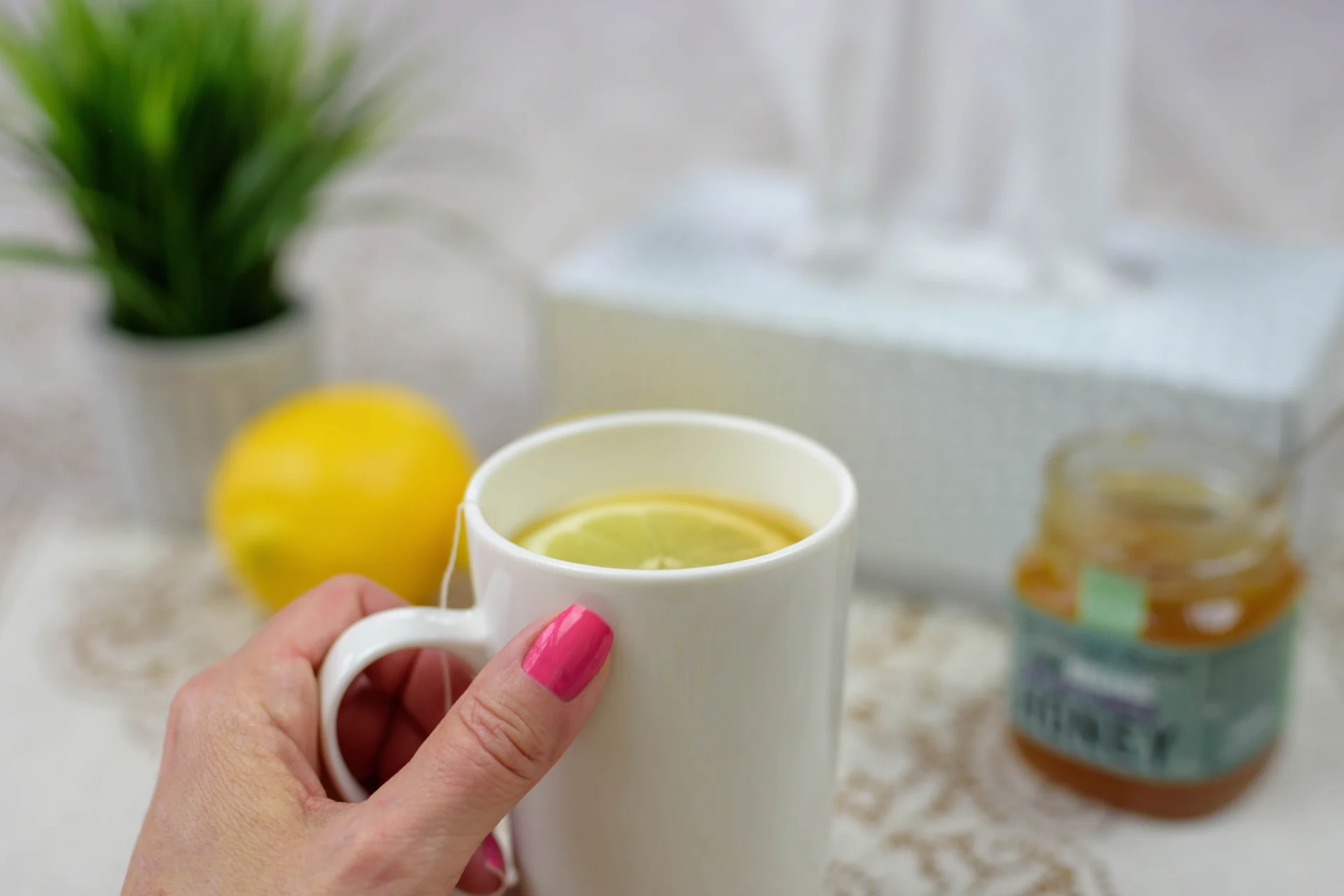 Hand hält eine Tasse Tee mit Zitronenscheibe, daneben Honigglas, Zitrone und Taschentücher – sanfte Hausmittel zum Halsschmerzen lindern